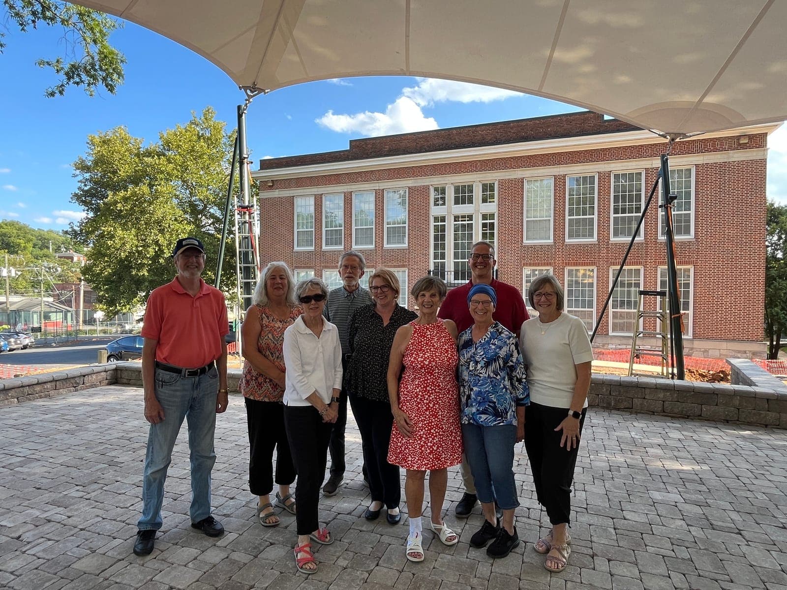A group of adults standing on the paved terrace under a white shade sail, with the brick Staunton Public Library building behind them