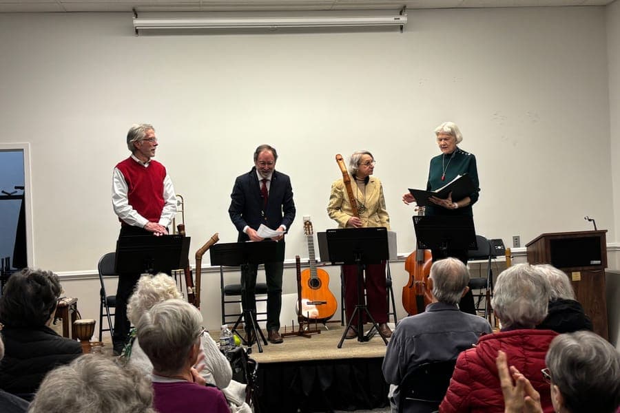 Four musicians standing behind music stands on a small stage in a library meeting room, with a guitar, recorders, and a viol beside them and a seated audience in the foreground